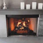 4 white candles on grey mantel above a burning gas log set with closed mesh curtain